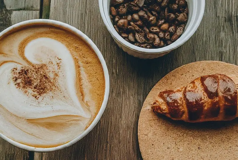 Taza de café con diseño de corazón, tazón de granos de café y un croissant sobre una mesa de madera.